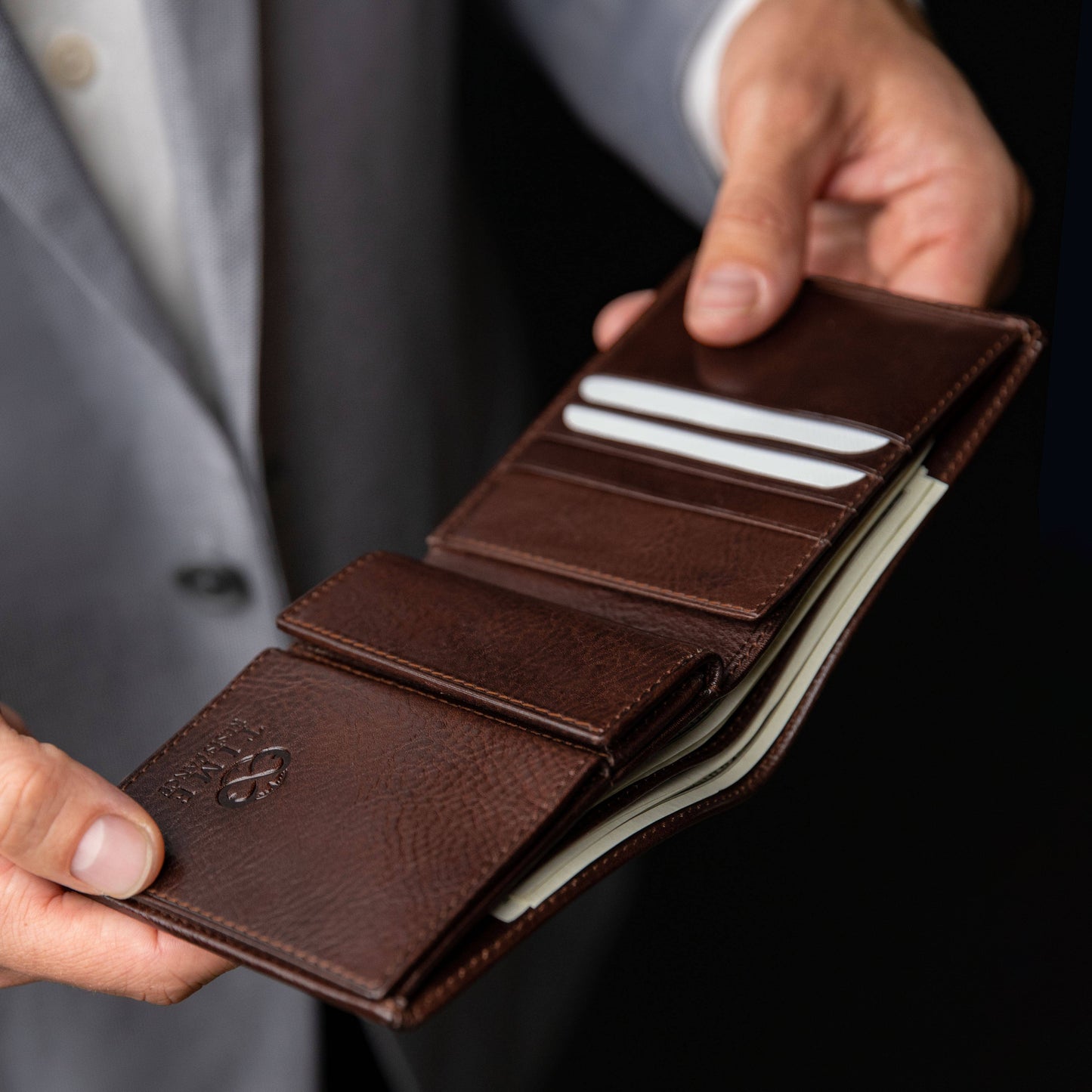 Brown leather wallet held by a person in a suit against a dark background