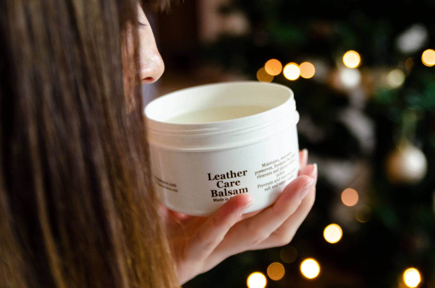 Person holding premium Leather Care Balm pot near Christmas tree with blurred lights.