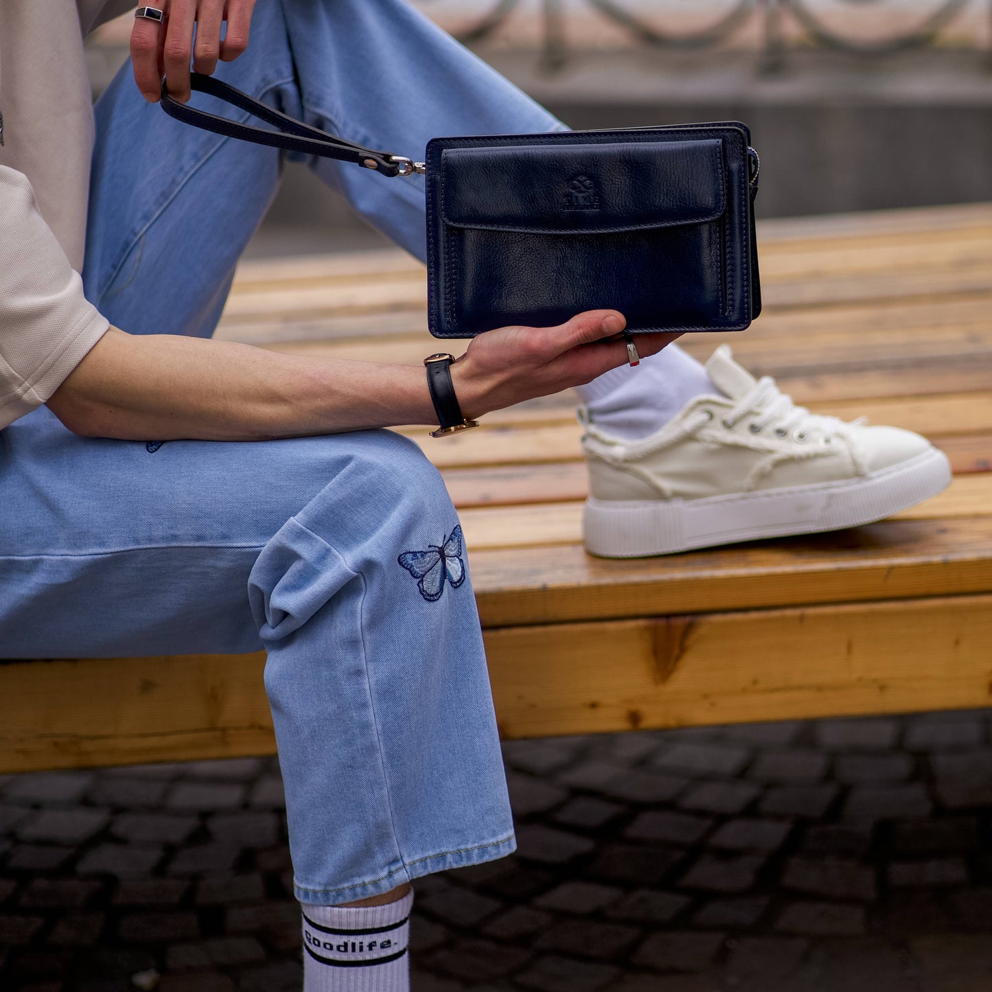 Person sitting on a bench holding a black clutch bag, wearing light blue jeans and white sneakers.