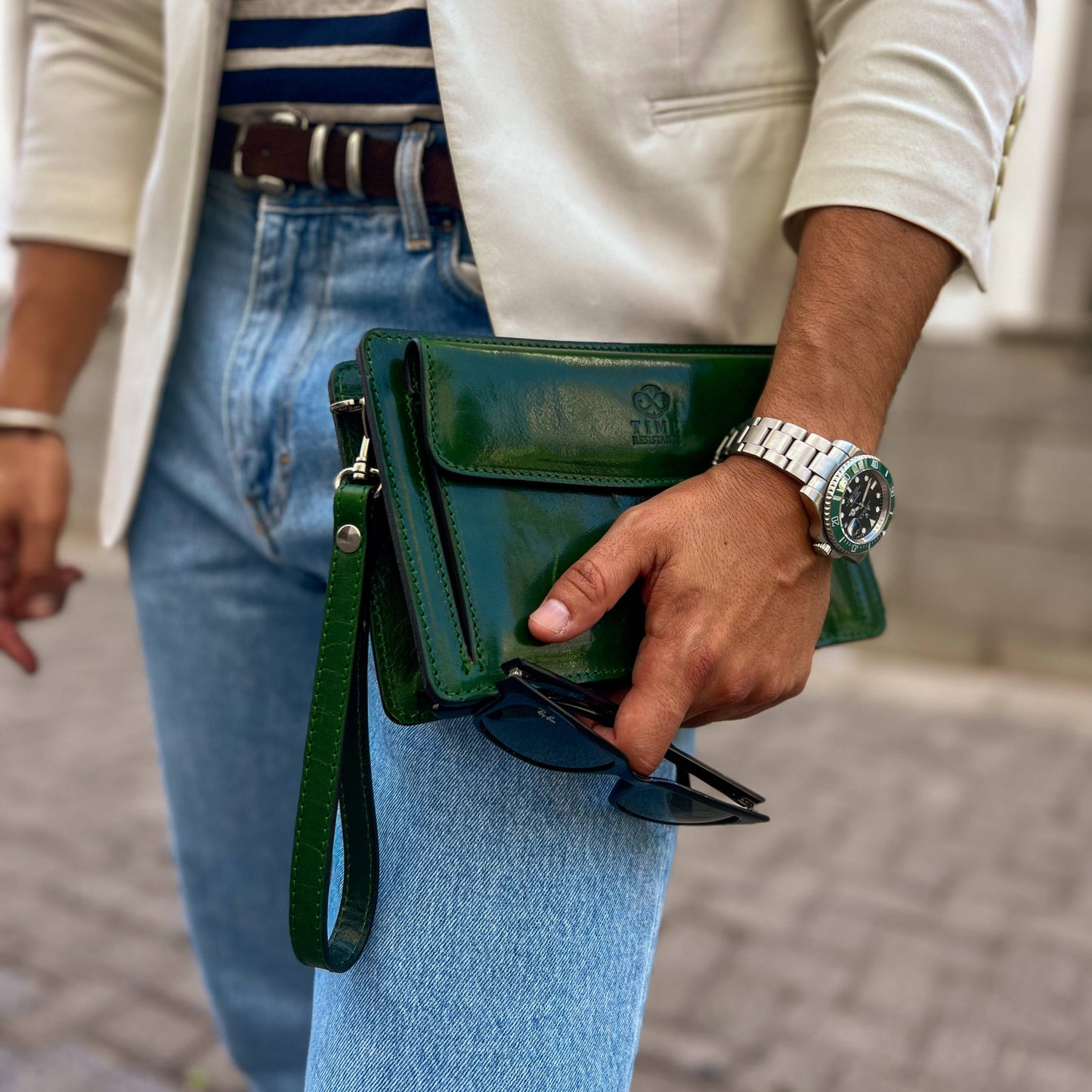 Person holding a green clutch bag with sunglasses, wearing a watch, on a blurred street background.