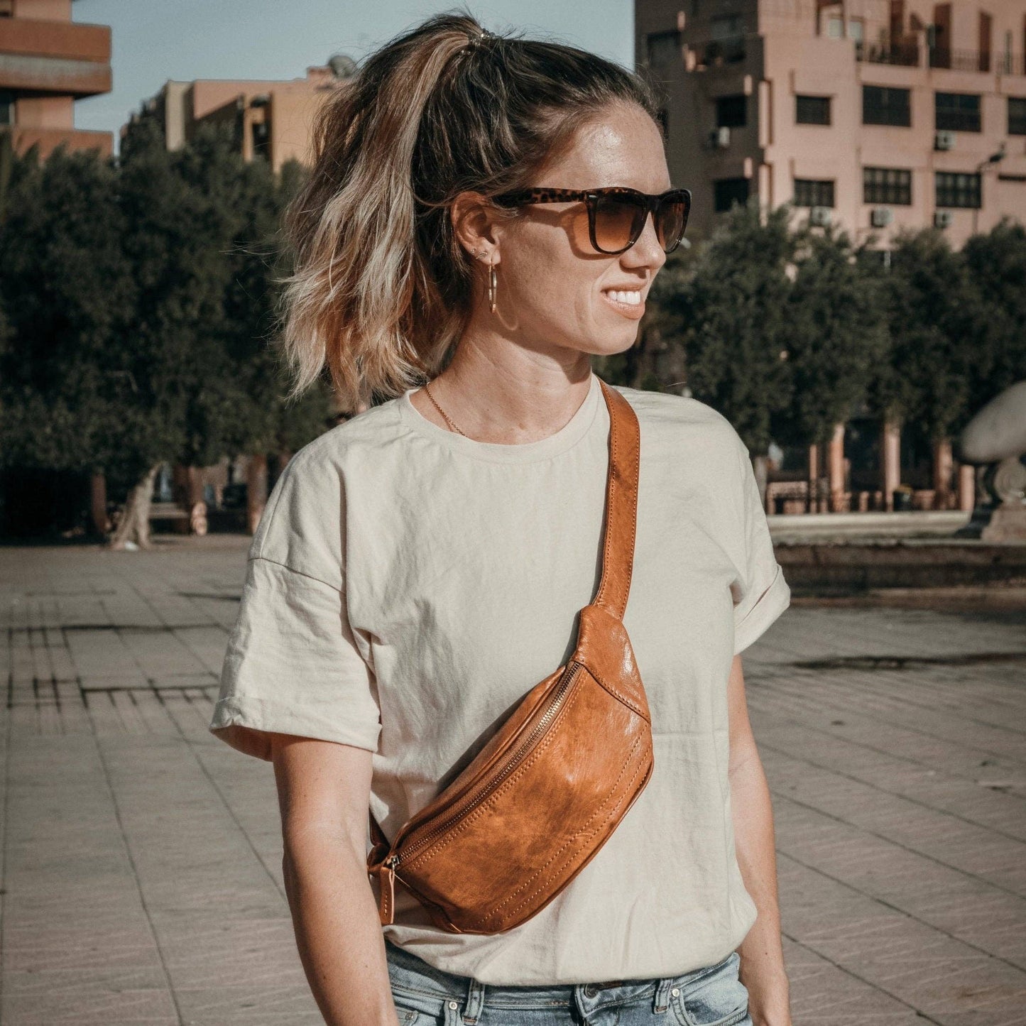 Woman wearing a cognac leather waist bag and sunglasses, standing outdoors in a casual outfit.