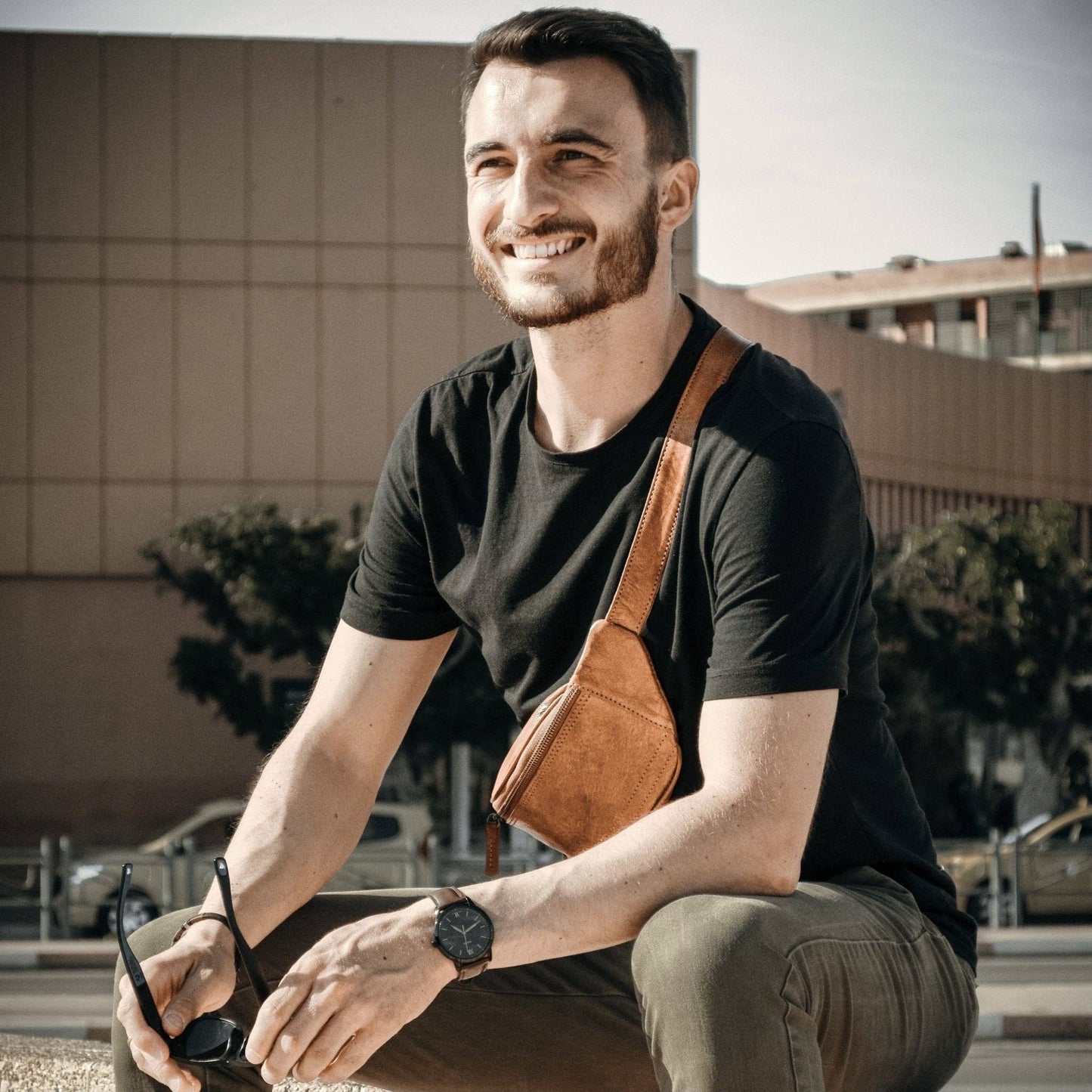 Man sitting outdoors wearing a black t-shirt, brown overalls, and holding sunglasses.