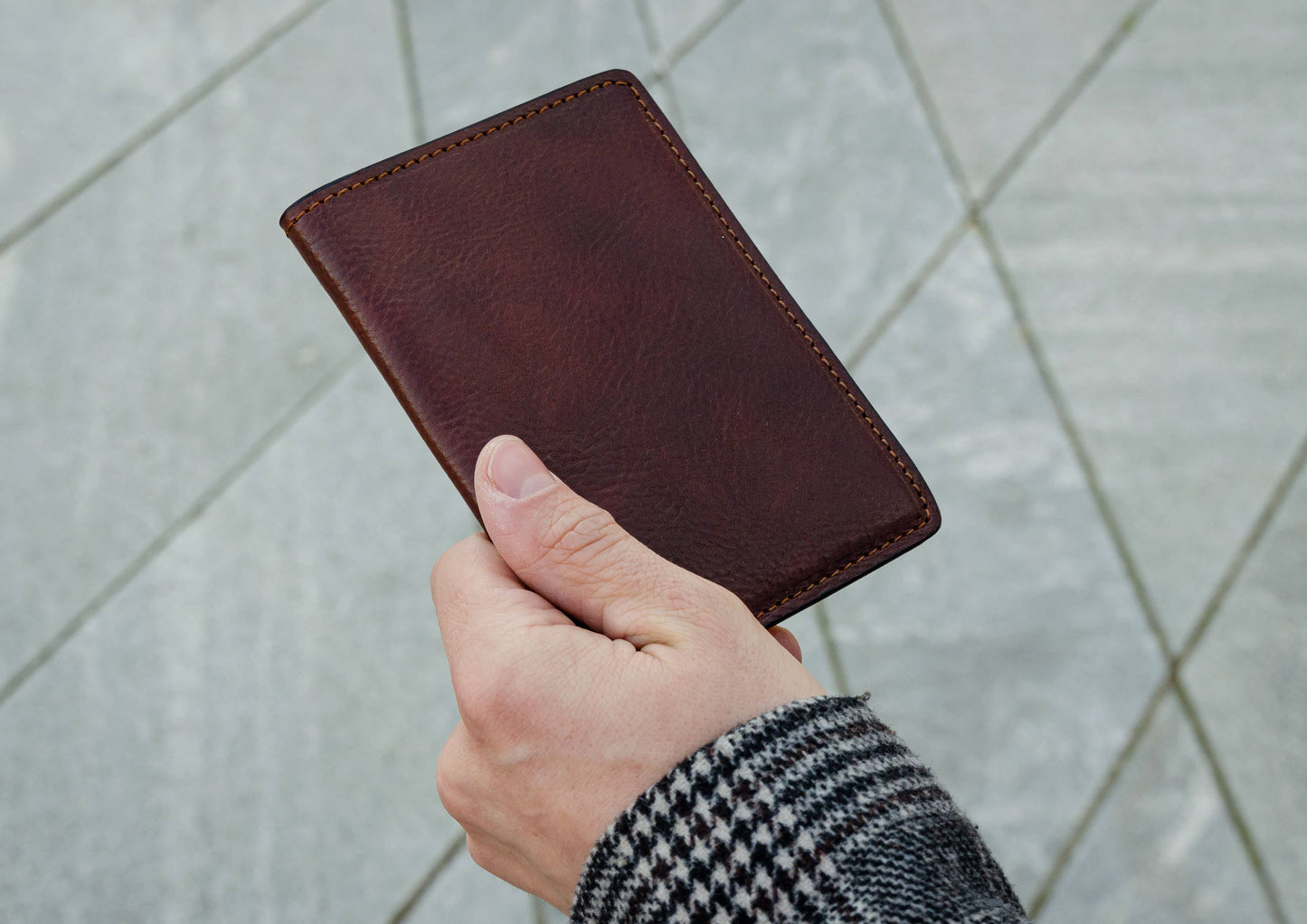 Hand holding a brown leather wallet against a light gray tiled wall.
