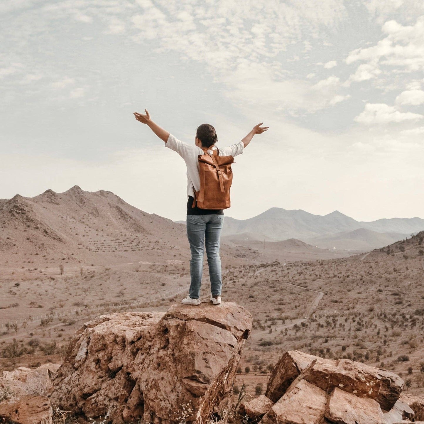 Person with a backpack standing on a rocky outcrop with arms outstretched, overlooking a desert landscape.