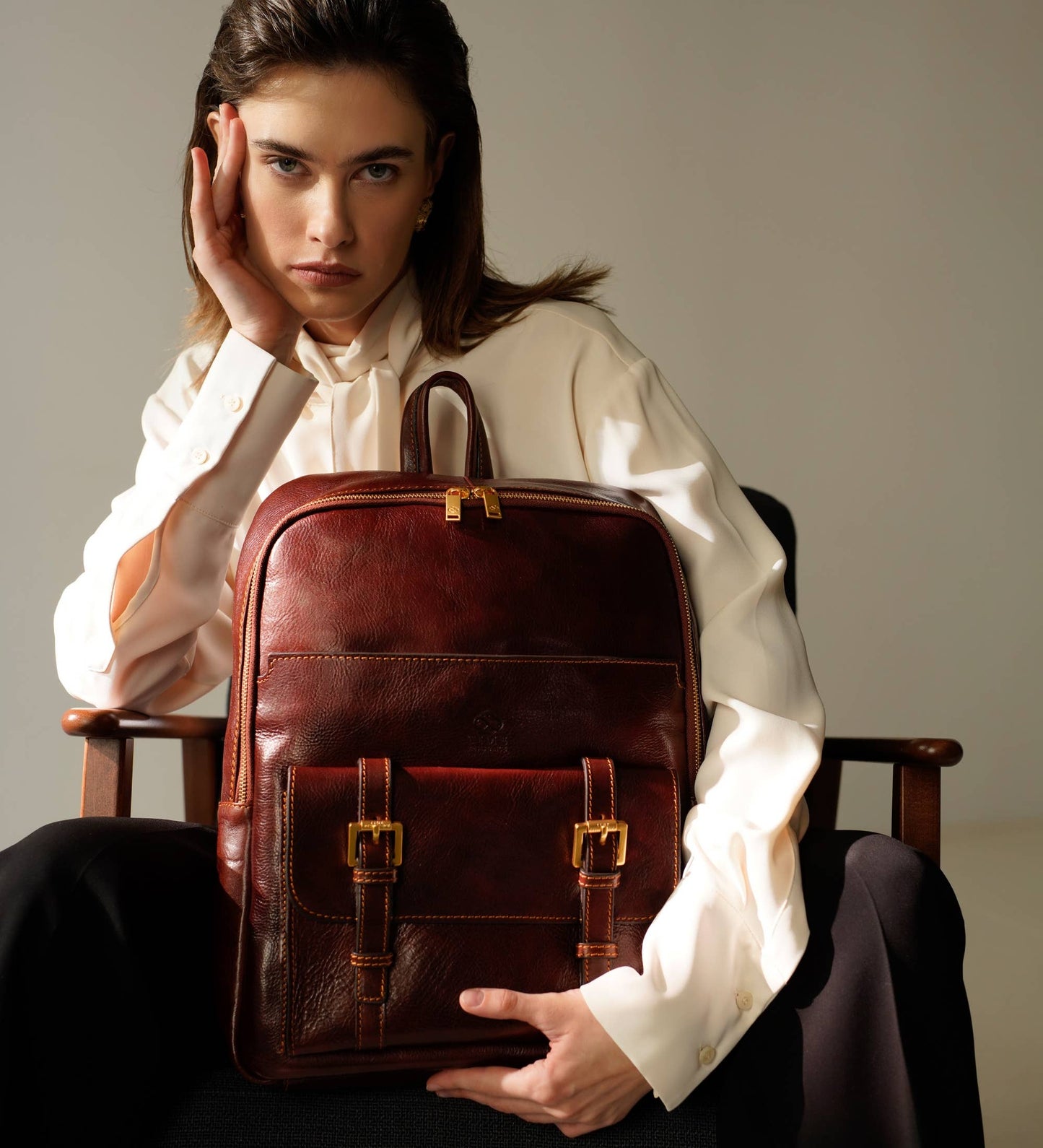 Woman holding a brown leather backpack in a studio setting