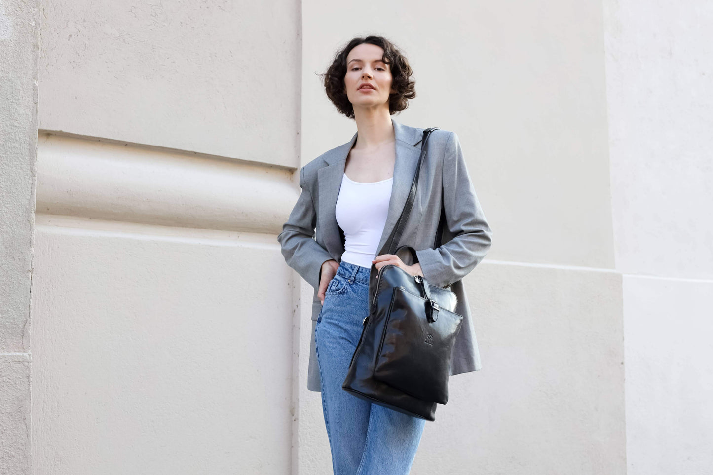 Woman in gray blazer and blue jeans holding a black bag against a light-colored wall.