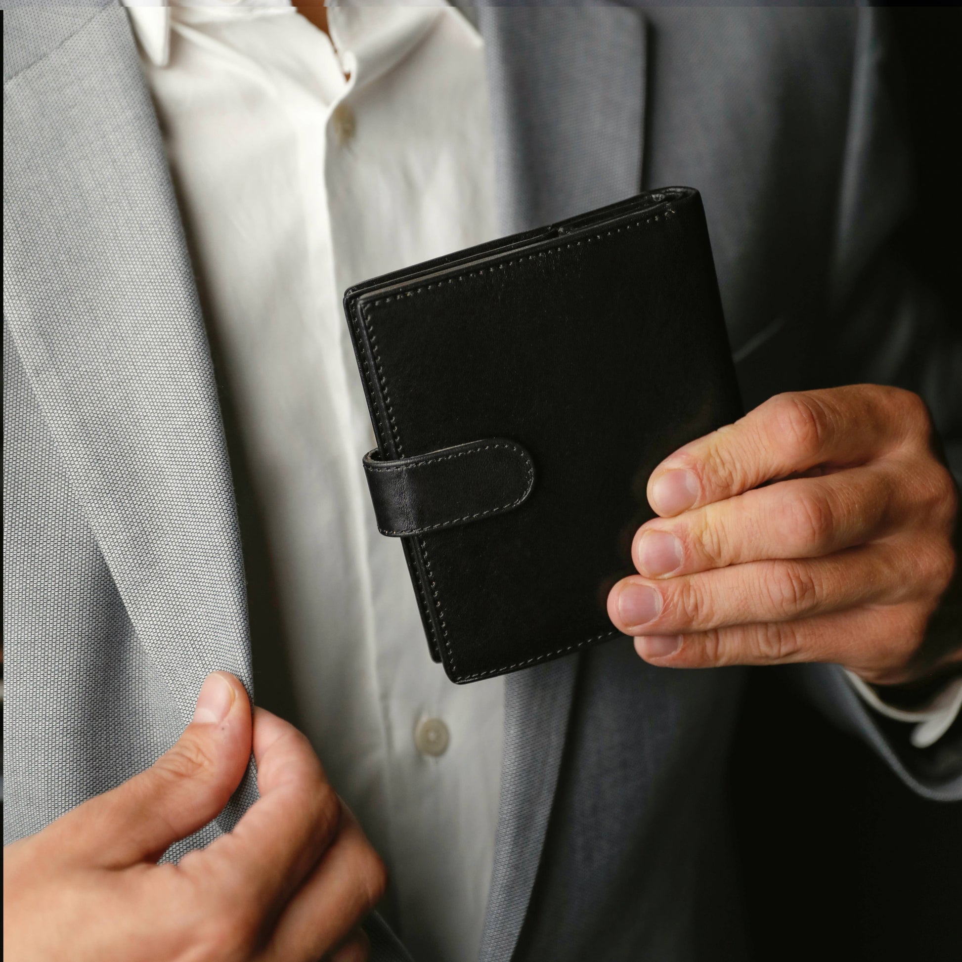 Man holding a sleek black leather bifold wallet with snap closure, dressed in a formal grey suit.