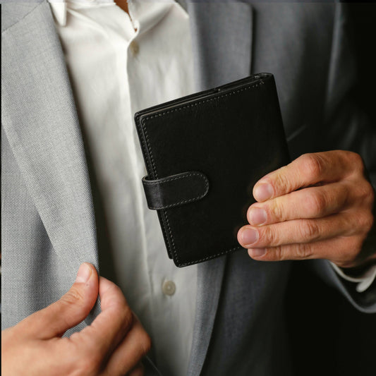 Man holding a sleek black leather bifold wallet with snap closure, dressed in a formal grey suit.