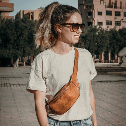 Woman wearing a cognac leather waist bag and sunglasses, standing outdoors in a casual outfit.