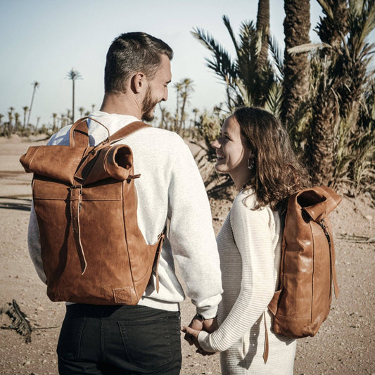 Two people with brown leather backpacks standing in a desert-like setting with palm trees.