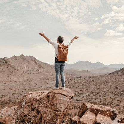 Person with a backpack standing on a rocky outcrop with arms outstretched, overlooking a desert landscape.