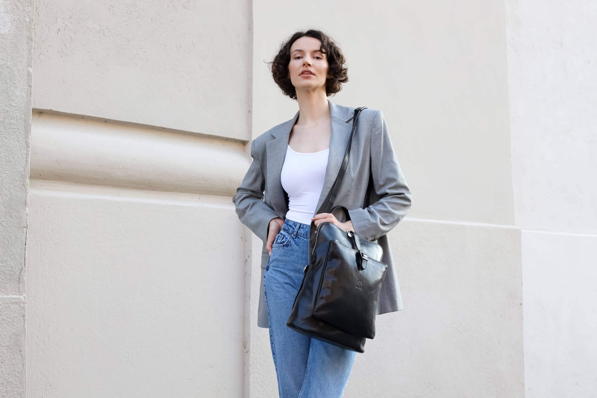 Woman in gray blazer and blue jeans holding a black bag against a light-colored wall.